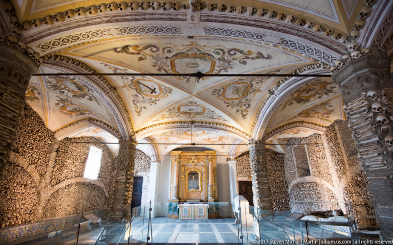 Axial view through Évora's Chapel of Bones toward a gilded altar, with bone-covered walls, piers, painted vaults, and glass barriers.