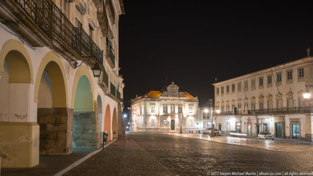 Night view across an Évora square with arcades at left and the illuminated Banco de Portugal building beyond.