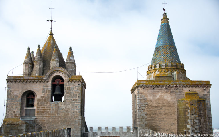 Square bell tower with visible bells beside a tall patterned conical spire on Évora Cathedral's rooftop.