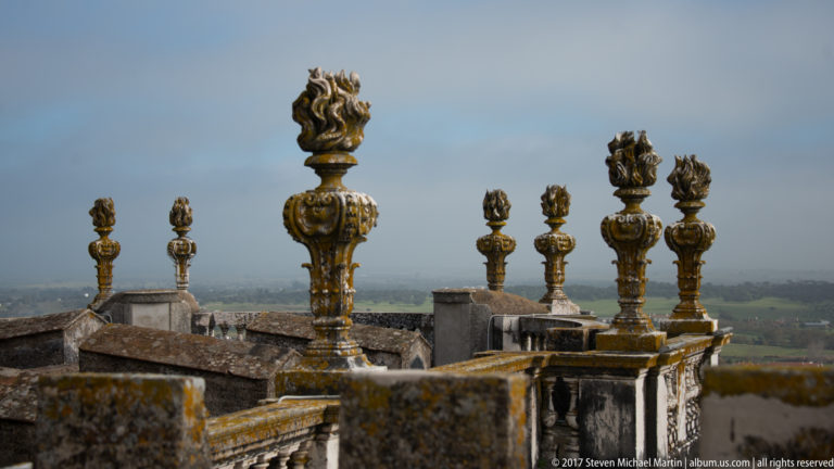 Row of weathered flame-shaped stone finials receding along Évora Cathedral's rooftop terrace toward the distant landscape.