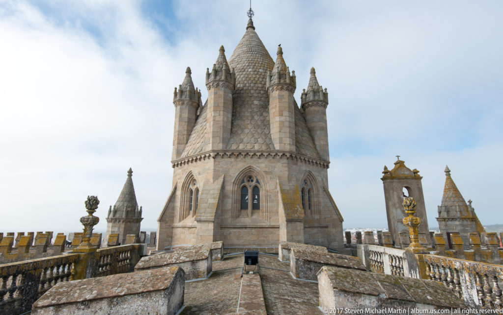 Large polygonal tower of Évora Cathedral with conical roof and corner turrets at the end of a crenellated rooftop walk.
