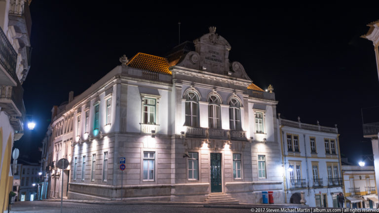 Illuminated corner view of Évora's Banco de Portugal building with arched windows, pediment, stone detailing, and tiled roof.