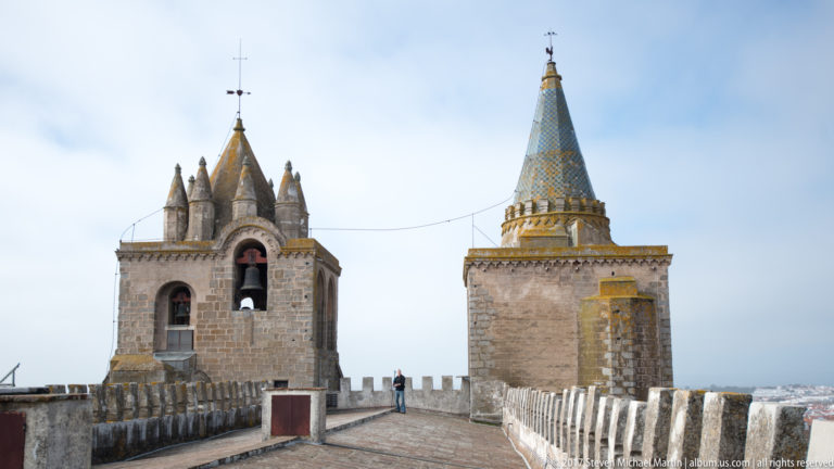 Bell tower and patterned conical spire rising from the crenellated rooftop terrace of Évora Cathedral.
