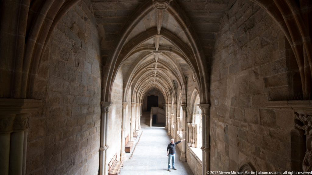 Long Gothic cloister corridor at Évora Cathedral with repeated pointed vaults and a lone visitor in the distance.