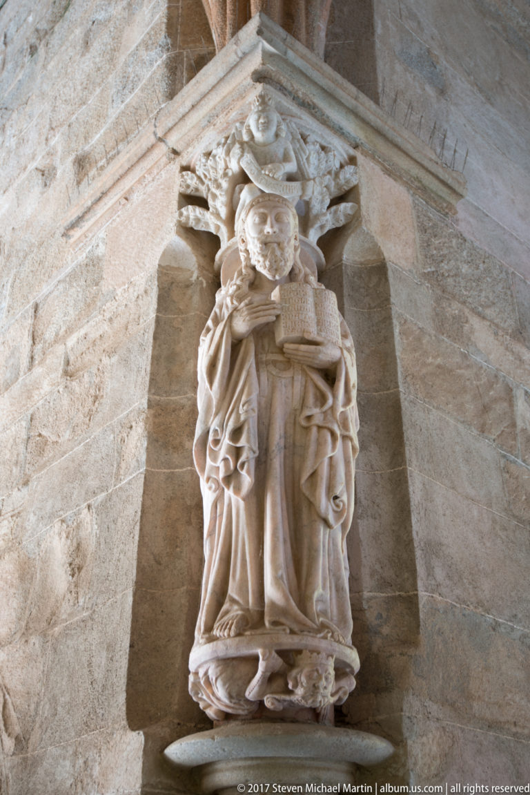 Carved stone robed cleric holding an open book beneath an ornate architectural canopy on the cathedral wall.