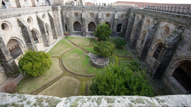 High view into Évora Cathedral cloister garden with paths, citrus trees, central stone feature, and surrounding Gothic arches.