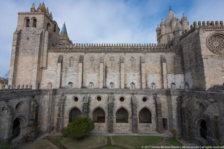 Évora Cathedral wall with buttresses and circular tracery window rising above the pointed arches of its cloister.