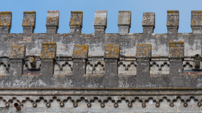 Weathered cathedral parapet with lichen-covered crenellations, small carved openings, and a repeating stone corbel course.