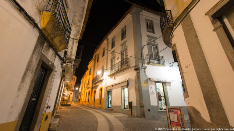Curving Évora street at night with whitewashed buildings, wrought-iron balconies, shopfronts, and warm lamps.