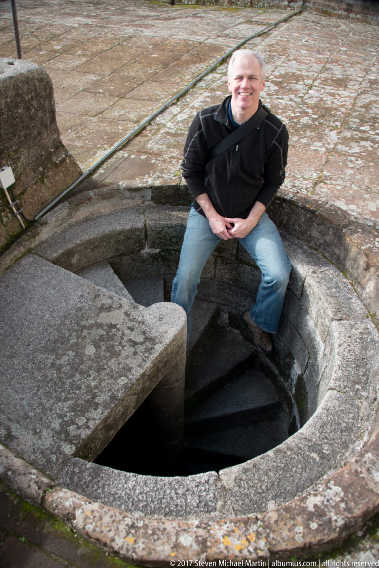 Visitor seated beside the circular opening of a worn stone spiral staircase on Évora Cathedral's rooftop.