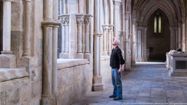 Visitor standing in Évora Cathedral's stone cloister beside clustered columns and pointed Gothic openings.
