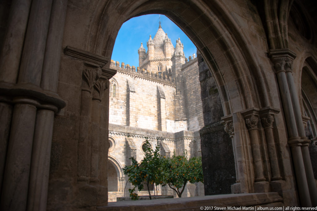 Évora Cathedral tower framed by a pointed cloister arch, with a citrus tree in the sunlit courtyard below.