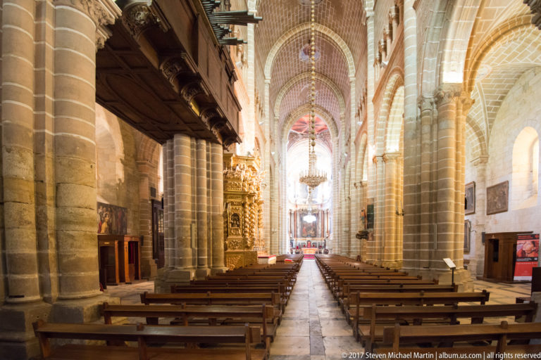 Rows of wooden pews extending through Évora Cathedral beneath ribbed vaults and between massive clustered stone piers.