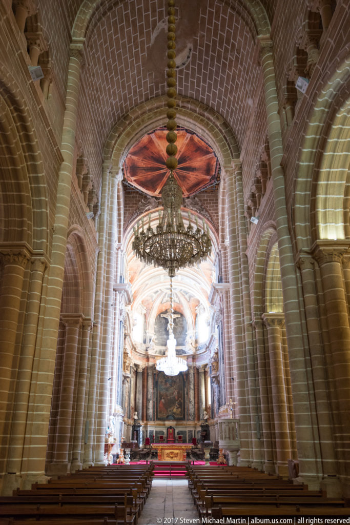Wide view down Évora Cathedral nave toward the sanctuary, framed by stone piers, pointed arches, vaults, and chandeliers.