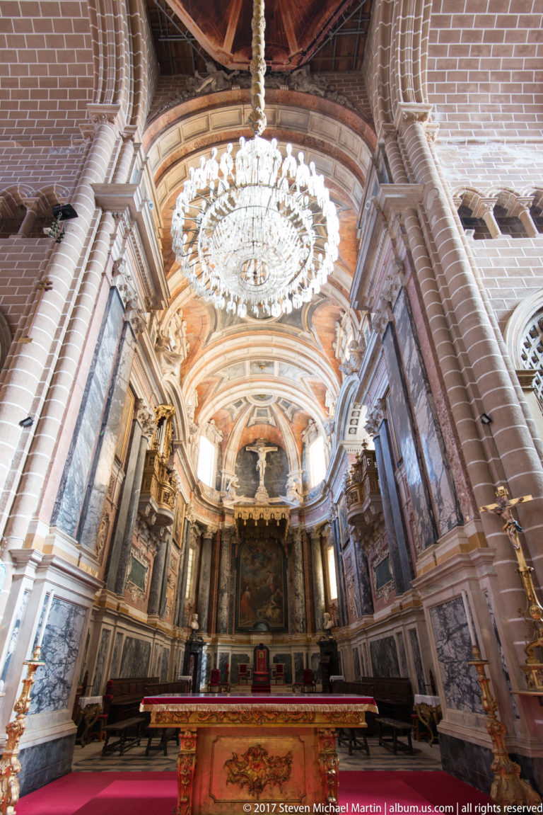Wide view of Évora Cathedral sanctuary with crystal chandelier, painted vault, marble wall decoration, sculpture, and altar.