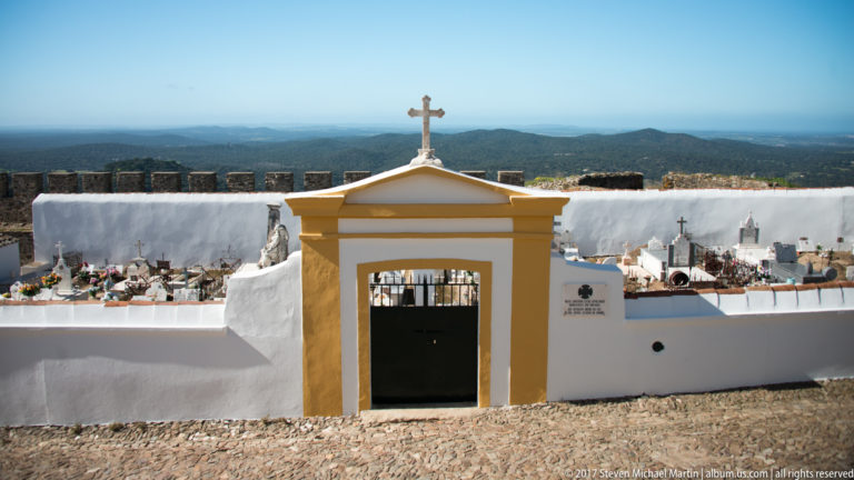 Yellow-trimmed white cemetery gate topped by a cross, with graves behind it and wooded hills extending into the distance.