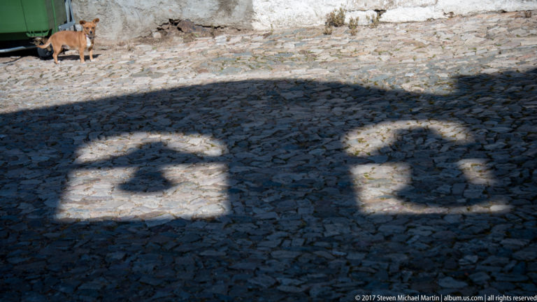 Two dark architectural shadows fall across rough gray cobblestones in bright sunlight.