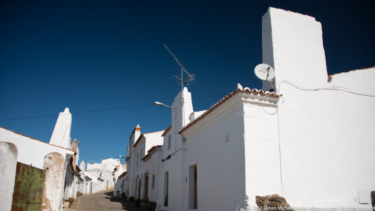 Narrow sunlit street lined with whitewashed houses, chimneys, tiled roofs, and antennas beneath a dark blue sky.