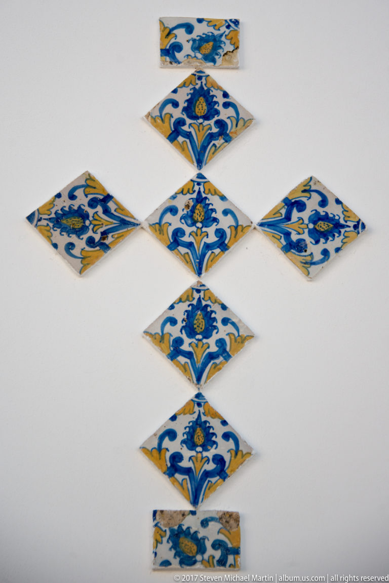 Blue, yellow, and white patterned ceramic tiles arranged in a cross-like design on a plain white wall.
