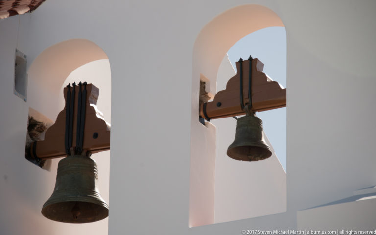 Two bronze bells suspended from wooden beams inside tall rounded openings in a whitewashed bell structure.