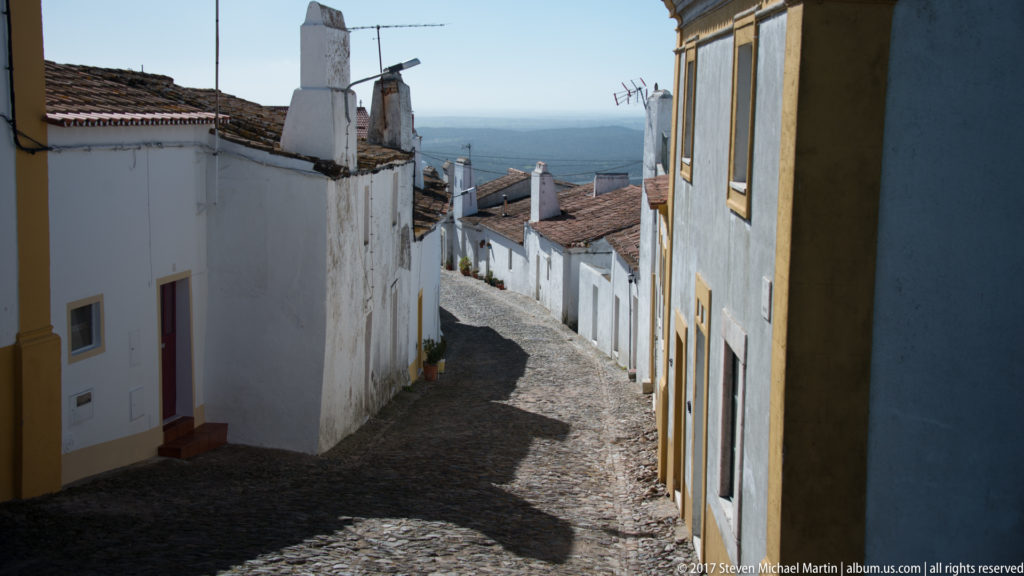 Steep cobbled lane descending between whitewashed houses toward a distant landscape, with ochre trim on the nearest façade.