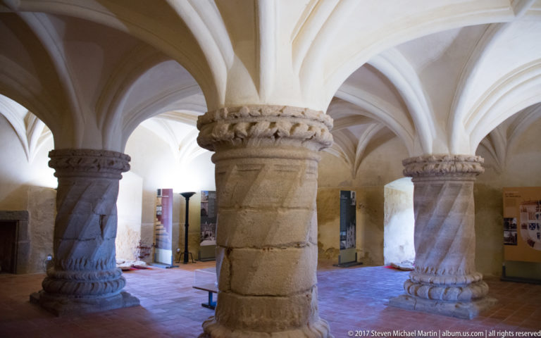 Three stone columns, including one spiral-carved shaft, support pale rib vaults above a terracotta floor inside the castle.