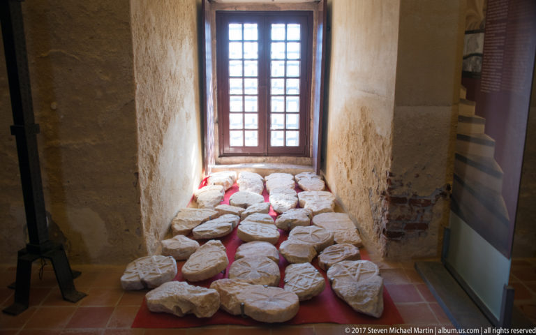 Pale carved stone fragments arranged on a terracotta floor beneath a tall gridded window inside a thick-walled room.