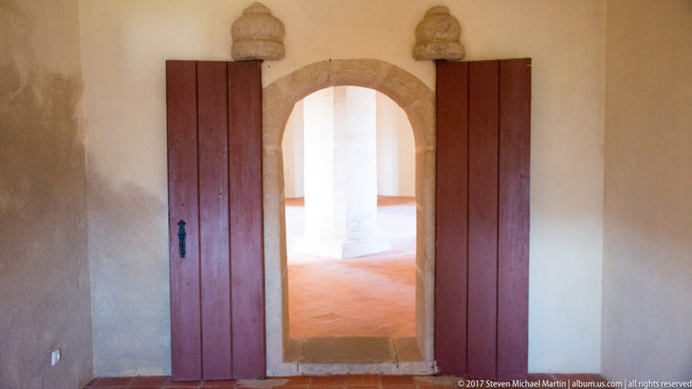 Arched stone doorway between reddish wooden leaves opening into a brighter castle room with terracotta floor and central pier.