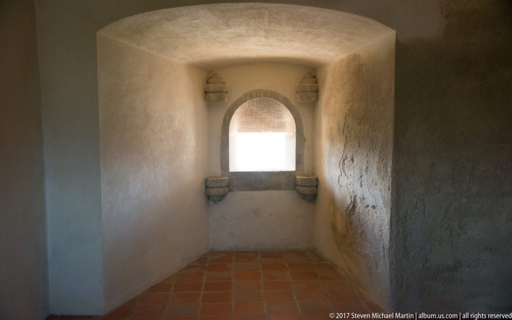 Deep arched window recess with side stone ledges in a thick plastered castle wall above terracotta floor tiles.