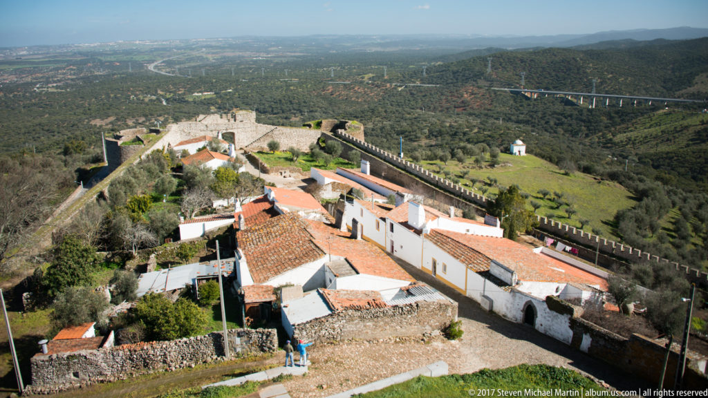 High view over red-roofed white houses enclosed by crenellated stone walls, with wooded countryside and a distant viaduct beyond.