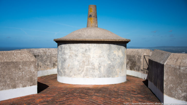 Round white rooftop structure with a conical stone cap and tall finial on a brick terrace behind low parapets.
