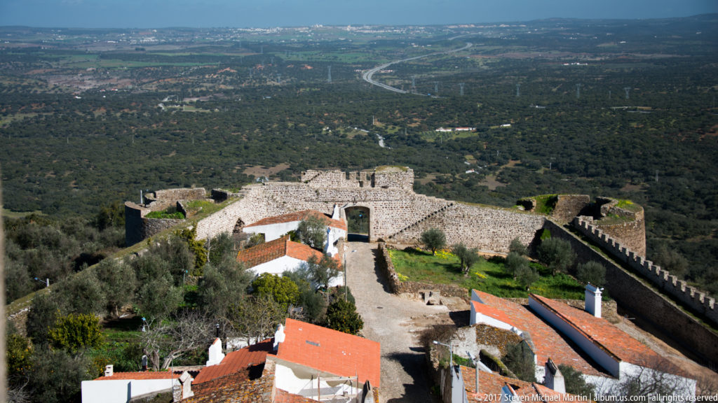 High view down onto a stone gateway, defensive walls, and red-roofed houses, with roads and countryside far beyond.