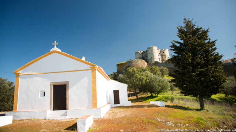 Small white chapel with ochre trim and a cross on its gable, with Evoramonte Castle rising on the hill behind.