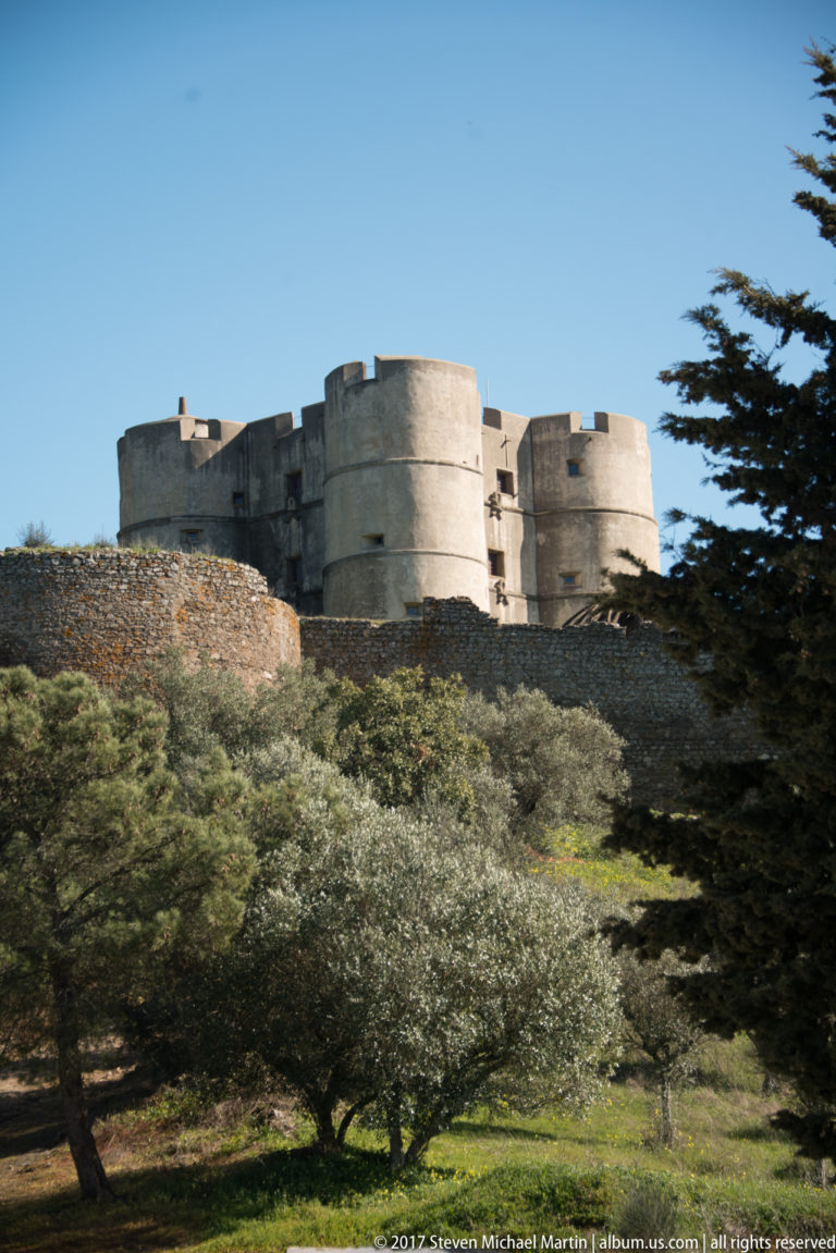 Castle of Evoramonte with four cylindrical corner towers rising above stone walls and olive trees under a clear blue sky.