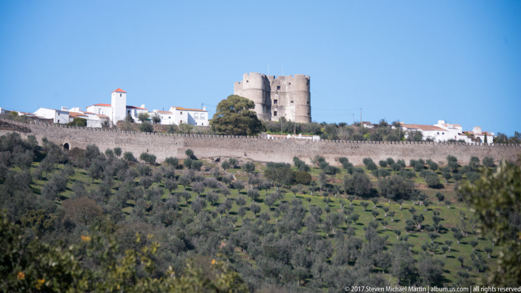 Four-towered Evoramonte Castle above white village buildings and a long stone defensive wall on a cultivated hillside.