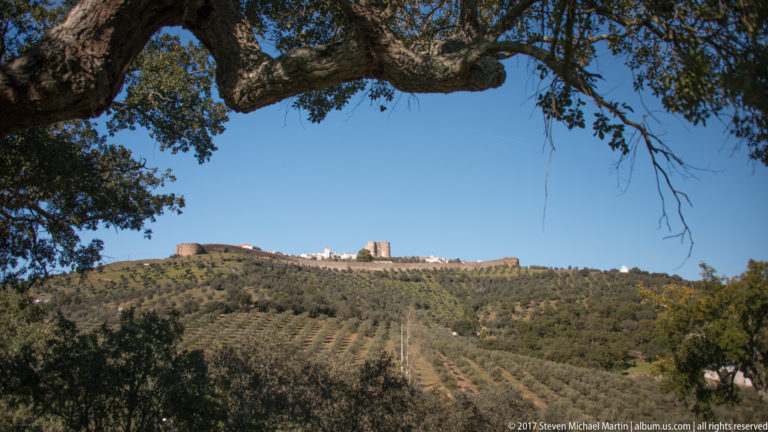 Distant fortified hilltop settlement and castle framed by cork-oak branches above patterned agricultural slopes.