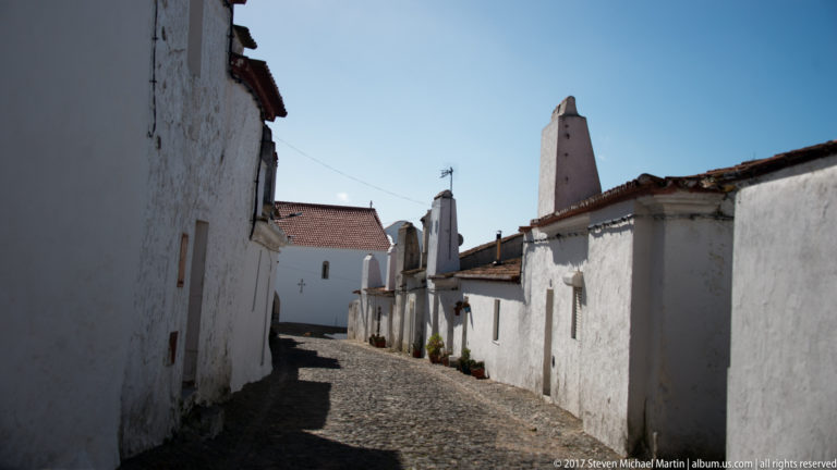 Narrow cobblestone lane curving between whitewashed houses and tiled roofs, with a tall vertical chimney-like structure ahead.