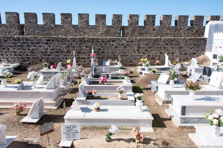 Rows of white graves decorated with flowers stand before a dark crenellated stone wall.
