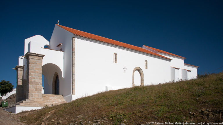 Whitewashed church with red tile roof, stone-framed side doorway, narrow windows, and a projecting arched entrance bay.