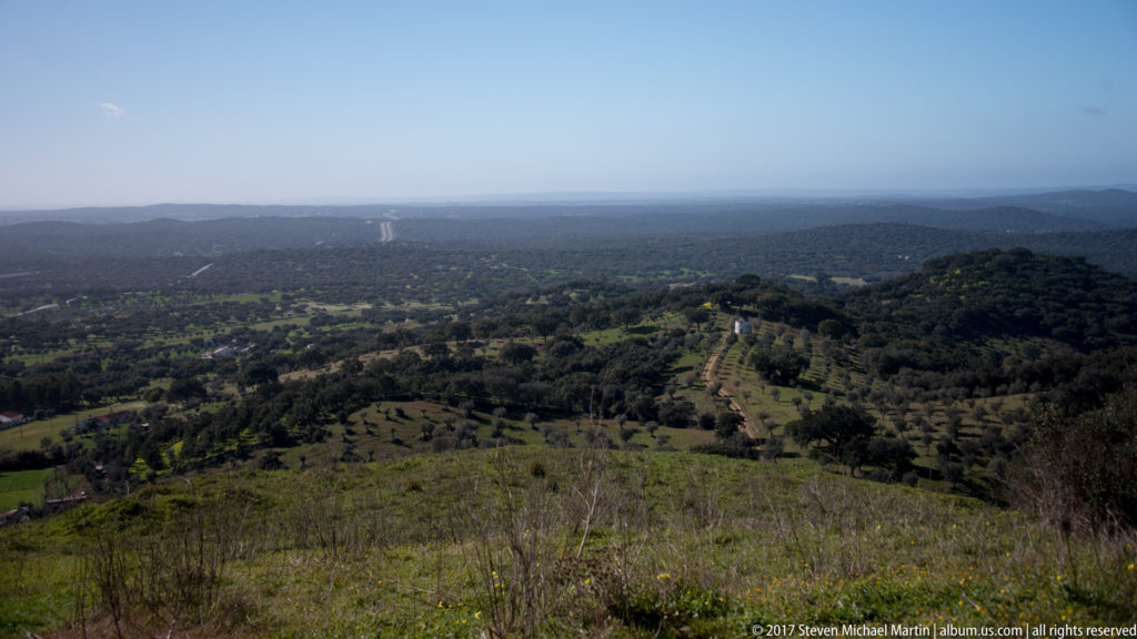 Wide elevated panorama of wooded hills, fields, tracks, and distant hazy plains below Evoramonte.
