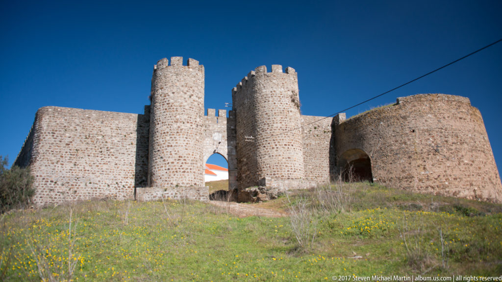 Stone gateway between two round crenellated towers in Evoramonte’s medieval walls, with grass and wildflowers in front.