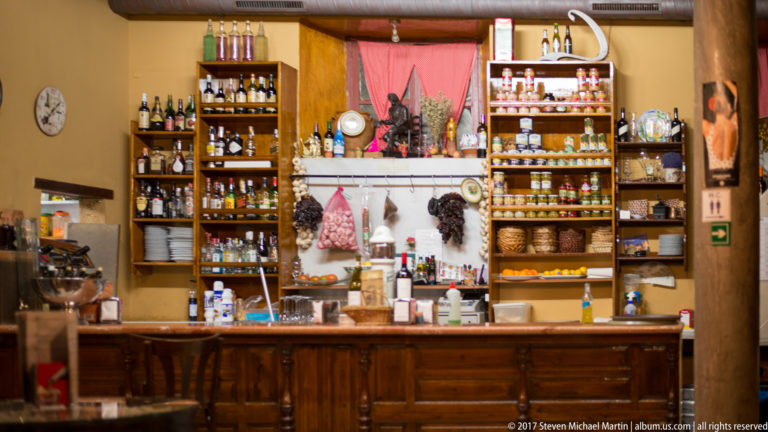Wooden bar counter with shelves of bottles, jars, cups, hanging cured meats, and a red fabric panel.