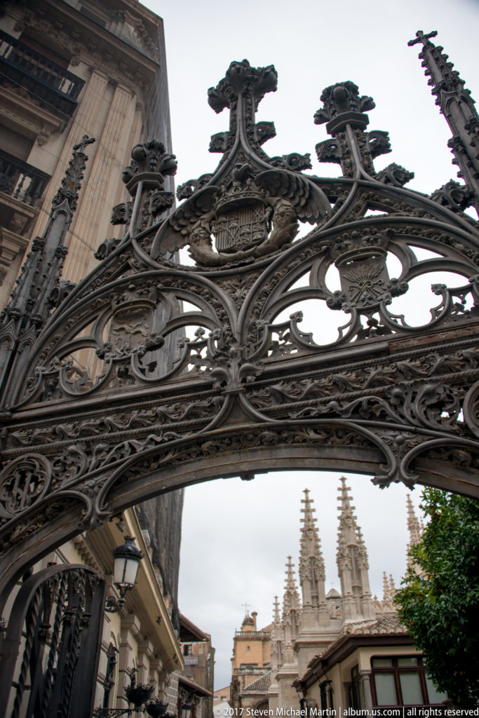 Ornate wrought-iron street arch framing the pinnacled upper architecture of Granada Cathedral under an overcast sky.