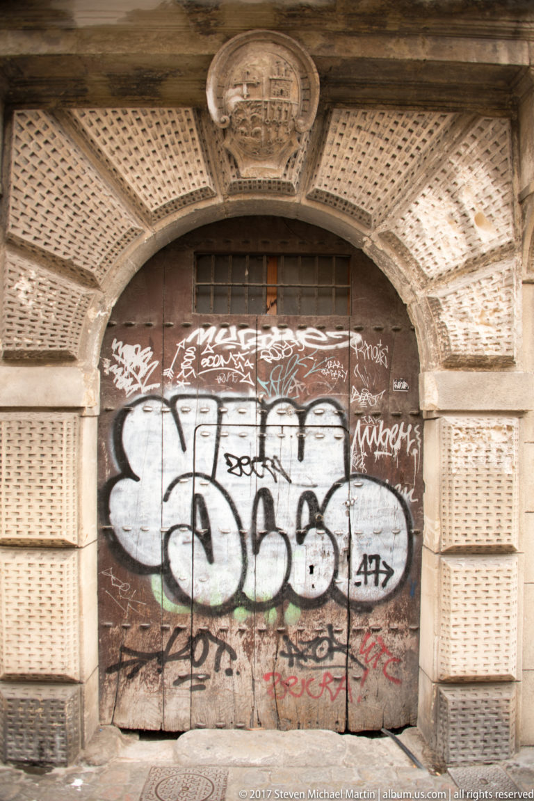 Graffiti-covered wooden double door beneath a carved semicircular stone arch and heraldic shield in Granada.