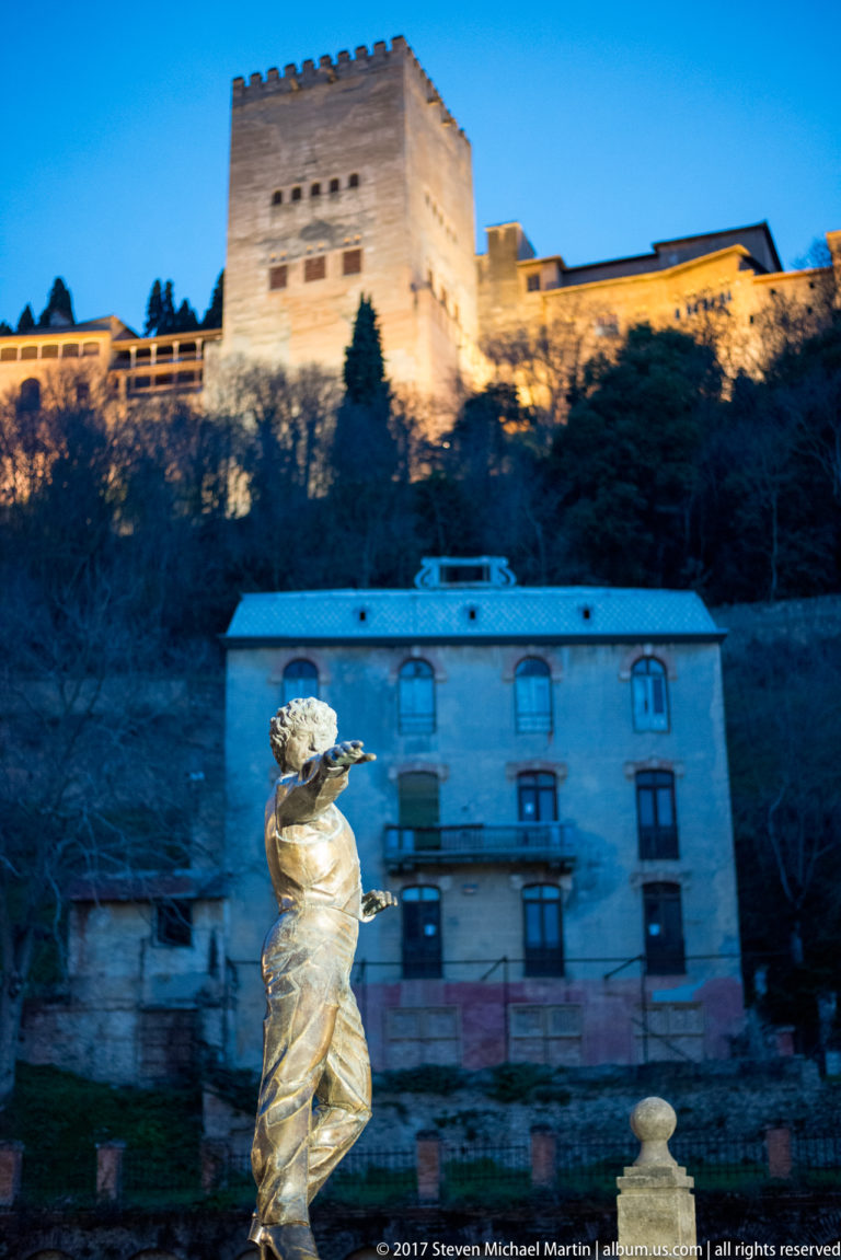Gilded female sculpture in the foreground with the illuminated Alhambra and square Torre de Comares behind.
