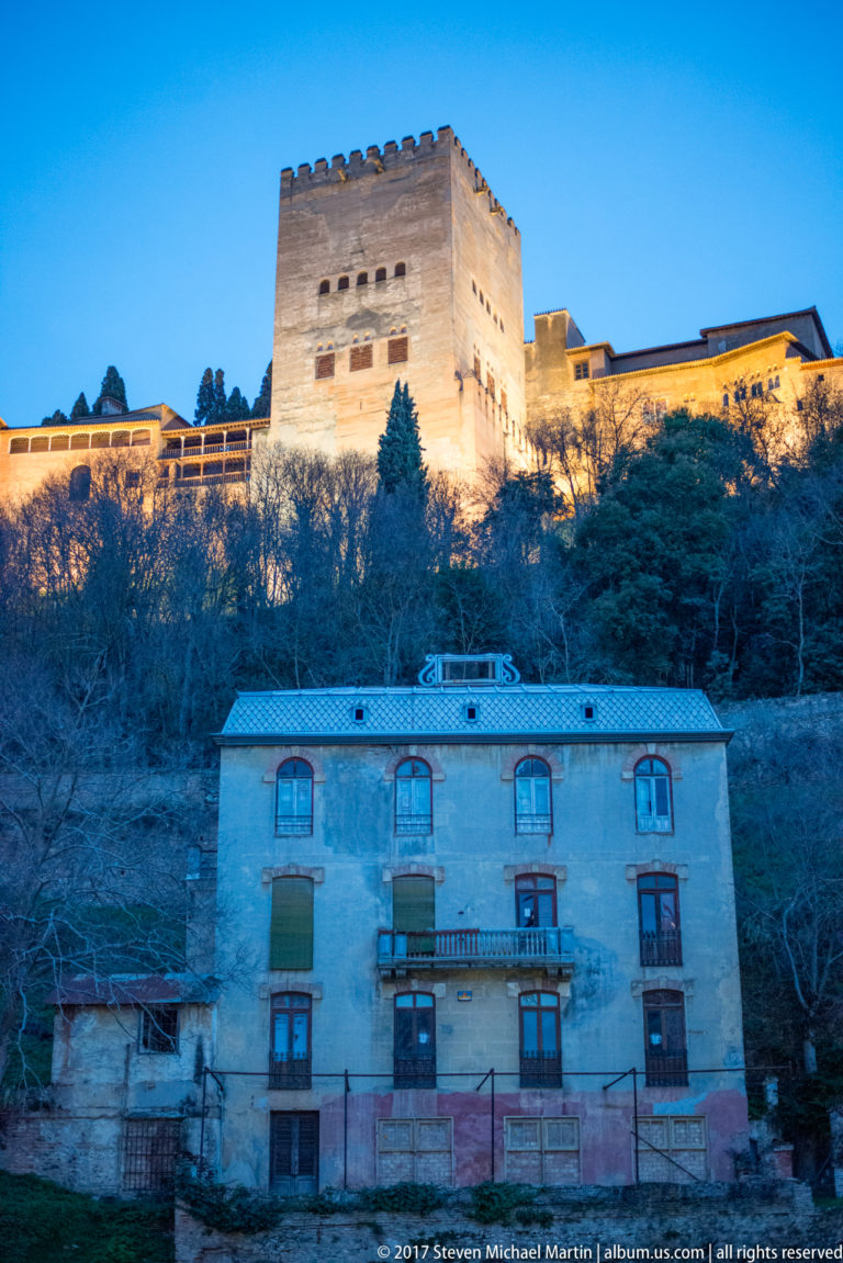 Illuminated Alhambra ridge with the square Torre de Comares above a residential building at dusk.