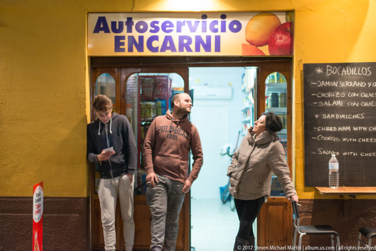 Three pedestrians outside a yellow takeaway shop with a chalkboard menu for bocadillos and sandwiches.