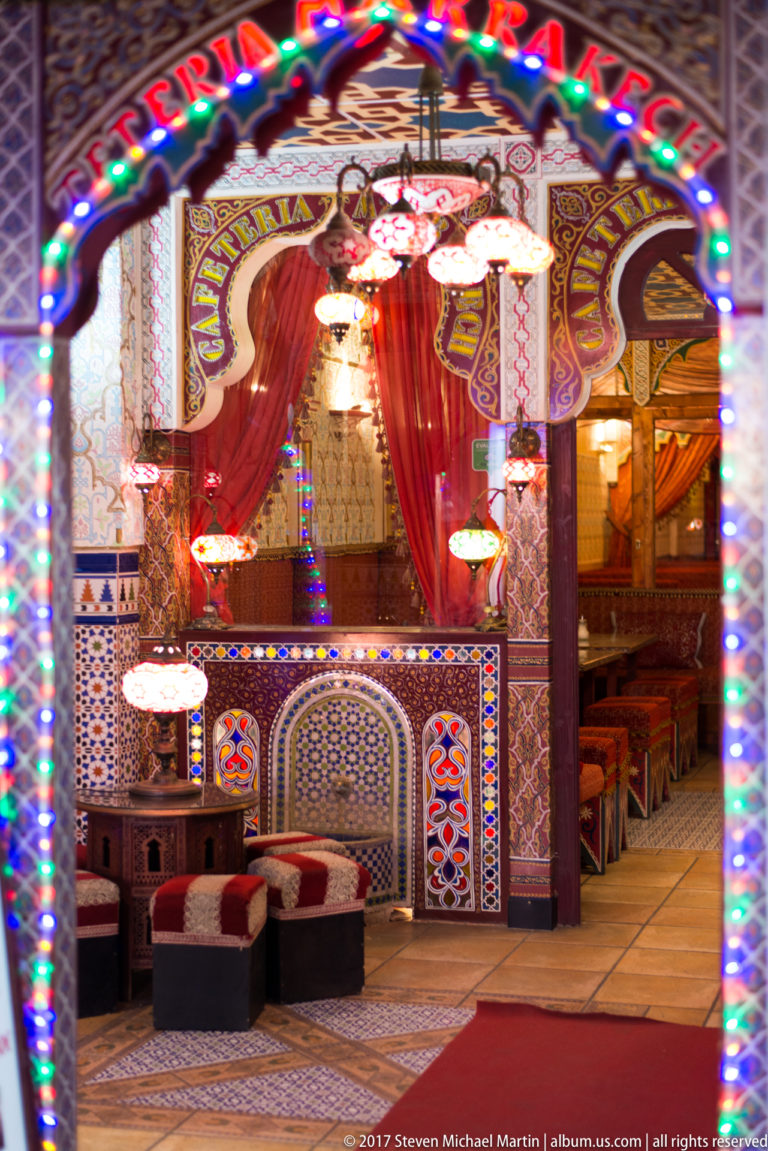 Ornate restaurant alcove with mosaic tilework, patterned arches, red drapery, hanging lamps, and low seating.