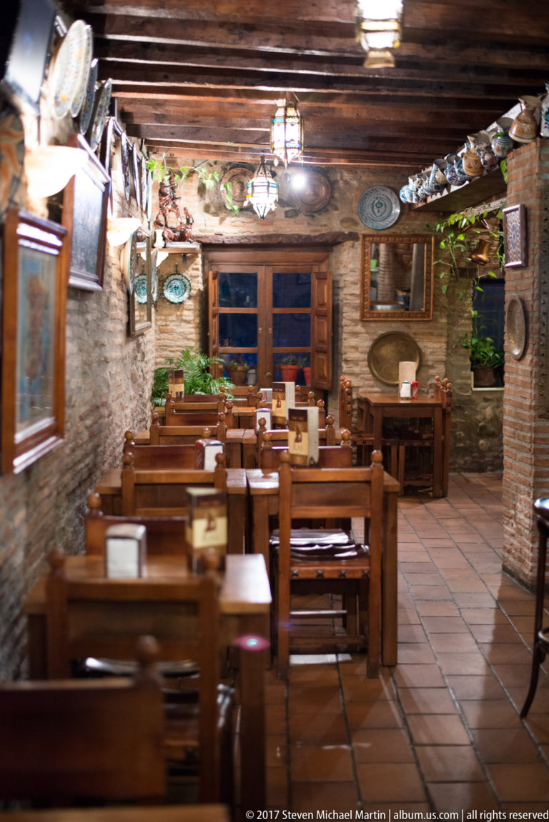 Empty rustic dining room with wooden tables and benches, timber ceiling beams, masonry walls, and hanging lanterns.