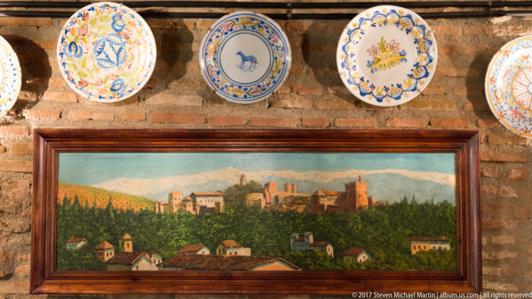 Framed painted view of the Alhambra beneath colorful ceramic plates on an exposed-brick wall.